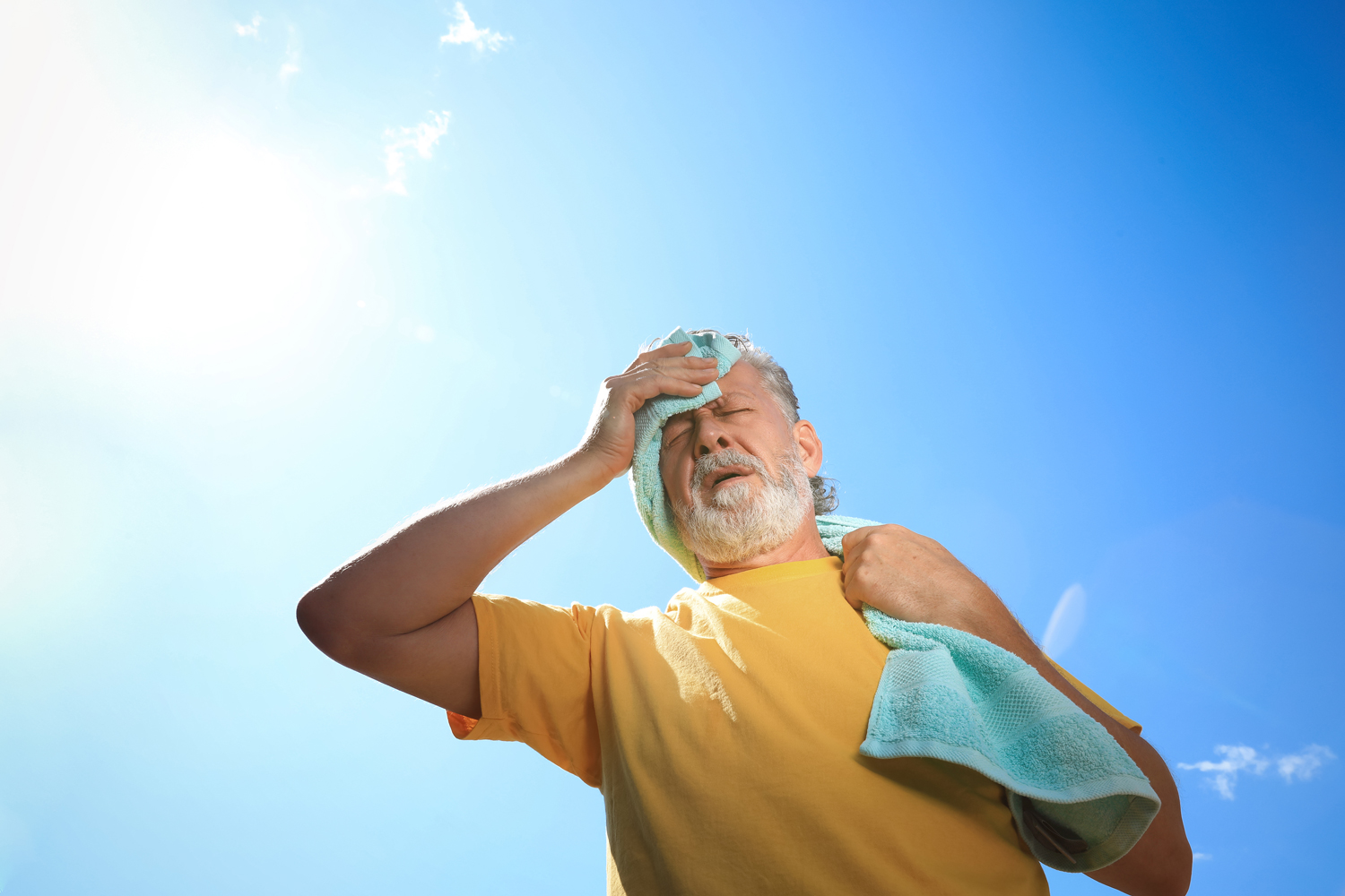 Ein Mann mit weiß-grauem Part, in gelbem Shirt, hält sich ein Handtuch an die Stirn. Von unten nach oben und gegen die Sonne fotografiert. Es ist viel blauer Himmel zu sehen.